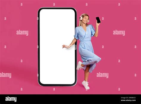 Happy Adult Caucasian Blonde Woman In Dress And Wireless Headphones Shows Smartphone Dancing