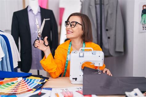 Middle Age Woman Tailor Using Sewing Machine Holding Scissors At Tailor