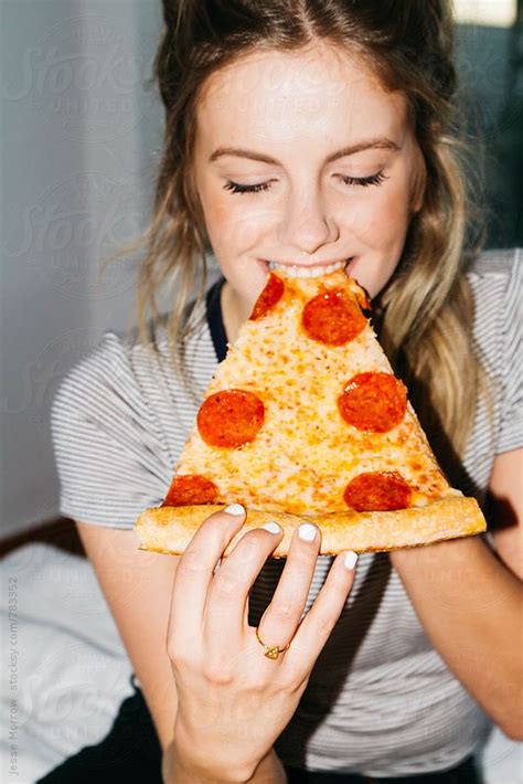 Young Woman Eating Slice Of Pepperoni Pizza On Bed In Apartment By