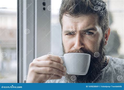 Grumpy Looking Bearded Man Drinking Morning Cup Of Coffee Near Window