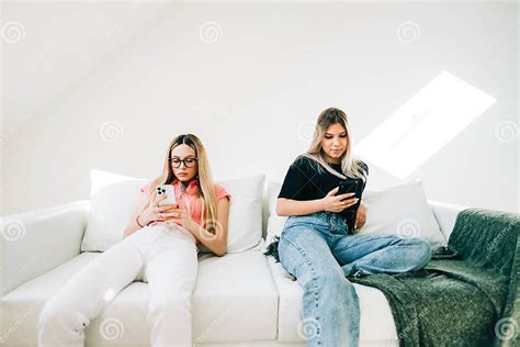 Two Women Sitting On Sofa In Living Room And Using Mobile Phones