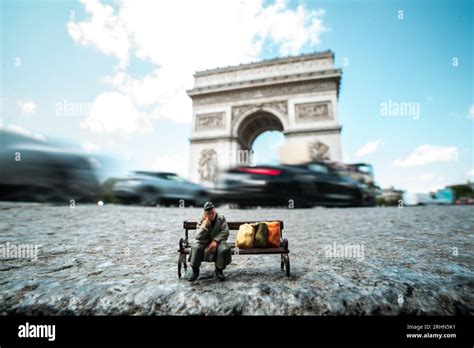 Miniature Homeless Man Sits On A Bench In Front Of The Arc De Triomphe