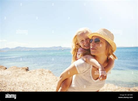 Lebhafte kleine blonde Mädchen mit ihrer Mutter am Strand lachen Reiten auf ihr in einem