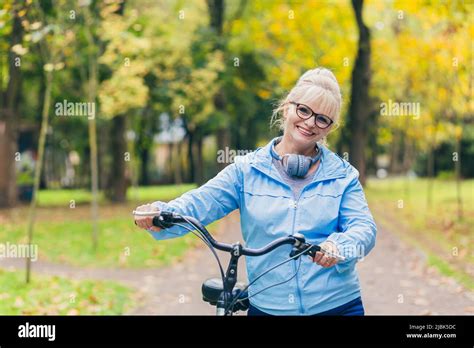 Senior Beautiful Blonde Woman Rides A Bike In The Park In The Woods In Nature Listens To