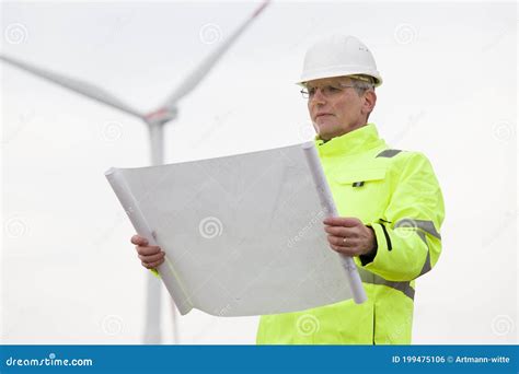 Mature Engineer Standing In Front Of A Wind Turbine With Blueprint In