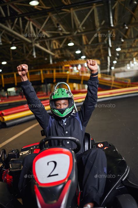 go kart speed drive, thrilled african american driver in helmet raising hands and winning kart ...