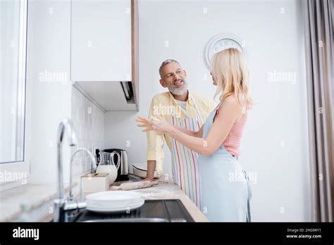 Positive Mature Man Talking To Wife With Dough In Kitchen Stock Photo