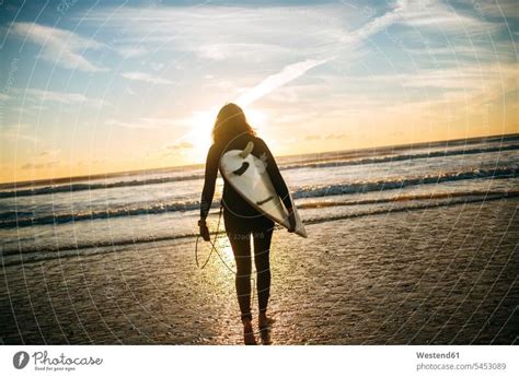 Back View Of Woman With Surfboard On The Beach By Sunset A Royalty