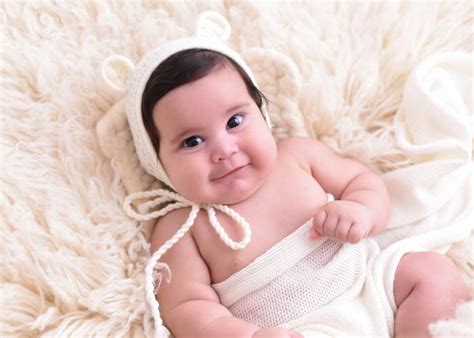 A Smiling Baby In A White Wrap Laying On A Fluffy Rug Wearing A Knitted Hat