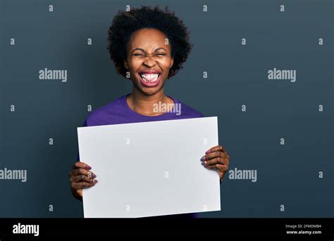 African American Woman With Afro Hair Holding Blank Empty Banner Sticking Tongue Out Happy With