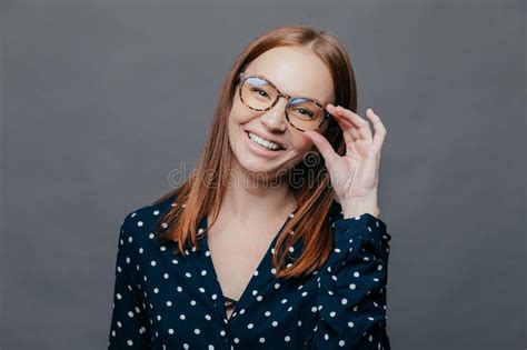 Smiling Woman With Pleased Facial Expression Keeps Hand On Rim Of Spectacles Wears Black Polka