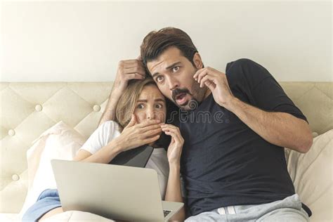 Scared Couple Sitting Under Blanket On The Bed With Laptop While