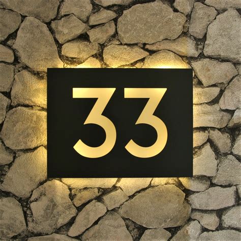 a black and gold house number sign mounted to a stone wall with lights ...