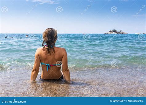 Atractiva Mujer En Bikini Sentada En La Playa Y Salpicada Por Las Olas Imagen De Archivo