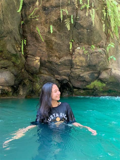 A Woman Is Floating In The Blue Water Near Some Rocks And Greenery