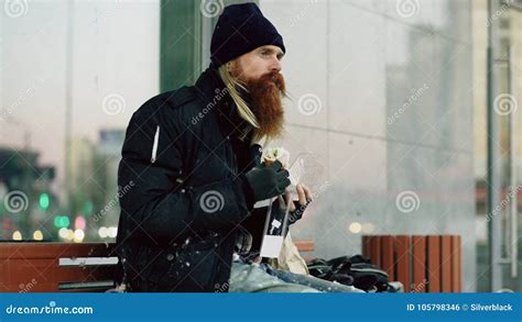 Homeless Young Man Eating Sandwich On Bench At City Street In Evening