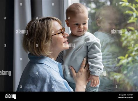 Side View Of Mature Blond Woman Smiling With A Baby Boy Near The Window