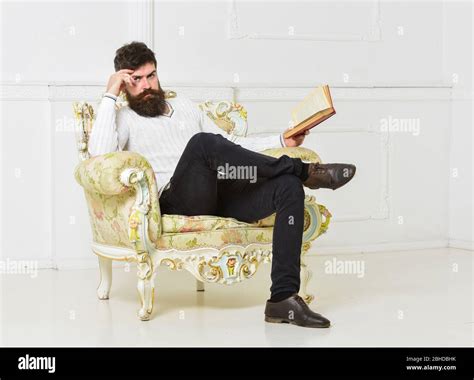 Man With Beard And Mustache Sits On Armchair Holds Book White Wall