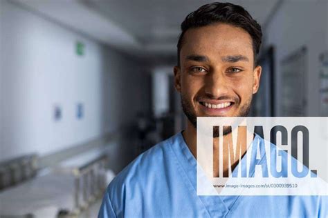 Portrait Of Happy Biracial Male Doctor Wearing Scrubs In Corridor At