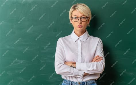 복사 공간이 칠판 앞에 닫힌 자세로 서 교실에서 안경을 쓰고 엄격한 젊은 금발의 여성 교사 무료 사진