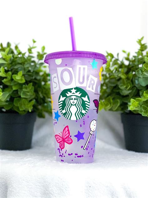 A Purple Starbucks Cup Sitting On Top Of A White Table Next To Potted