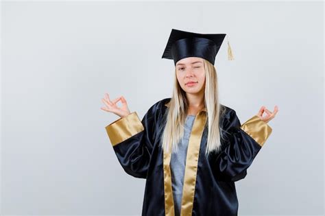 명상 포즈에 서있는 금발 소녀 졸업 가운과 모자에 윙크하고 행복을 찾고 무료 사진