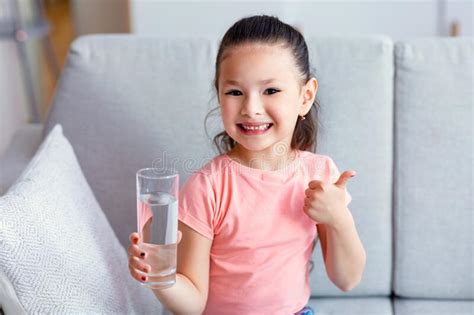 Chinese Girl Holding Glass Of Water Gesturing Thumbs Up Sitting Indoors