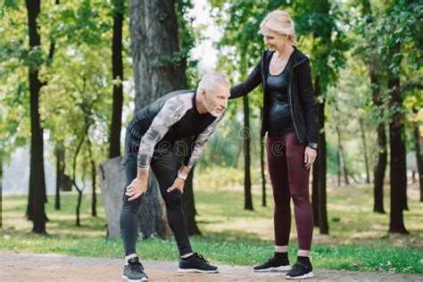 Sportswoman Standing Near Exhausted Mature Sportsman In Park Stock
