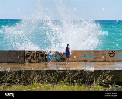 A Father And His Young Son Brave The Seawall As A Large Wave Crashes