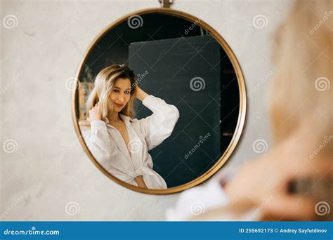 A Beautiful Blonde In A White Shirt Looks At Her Reflection In A Round