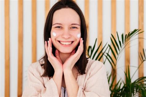 Premium Photo Smiling Young Woman Applying Cream On Face Looking At