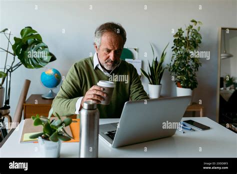 Mature Man Drinking Coffee While Working On Laptop At Home Stock Photo