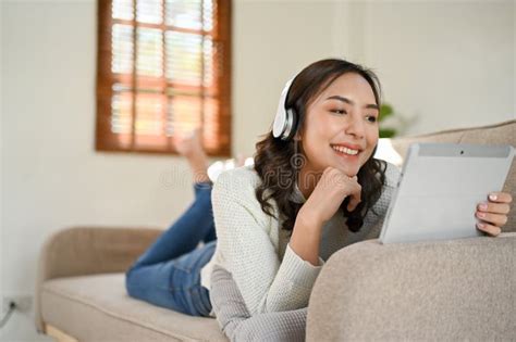 Attractive Asian Woman Lying On Sofa Wearing Headphones Watching