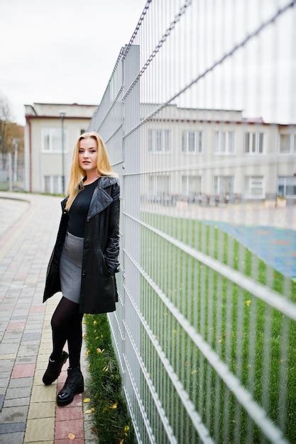 Fille Blonde à La Mode En Long Manteau De Cuir Noir Contre Une Clôture De Fer Dans La Rue