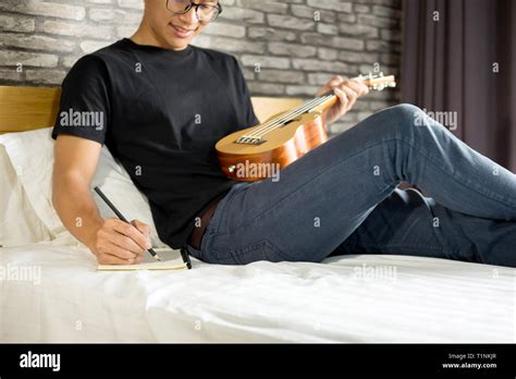 Happy Young Asian Man Playing Ukulele Sitting On Bed In Bedroom Stock