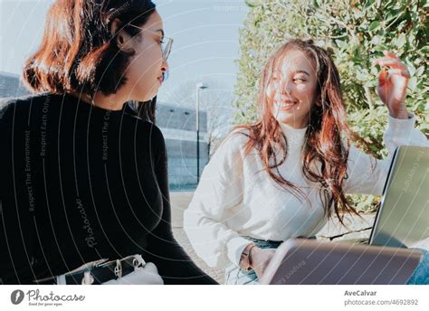 Two woman students talking and discussing a project while working on laptops at the university ...