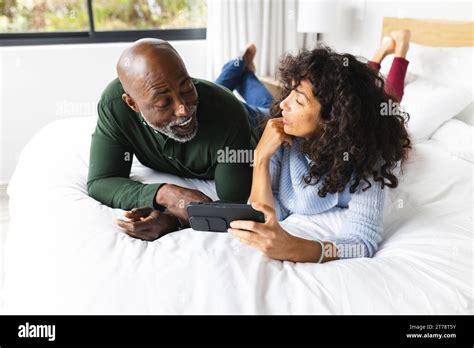 Happy Diverse Mature Couple Lying On Bed Using Smartphone Talking In