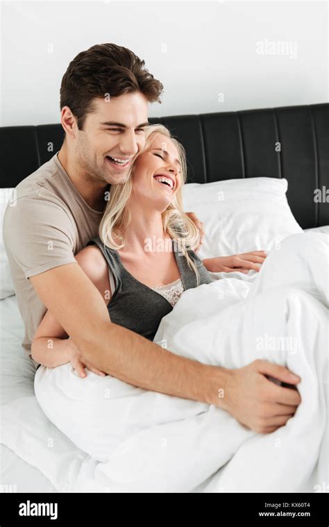 Happy Blonde Lady Laughing While Lying In Bed With Her Cheerful Brunette Man And Hugging Stock