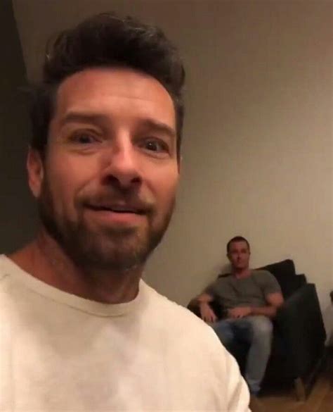 A Man In A White T Shirt Is Smiling At The Camera While Two Men Are Sitting On Couches Behind Him