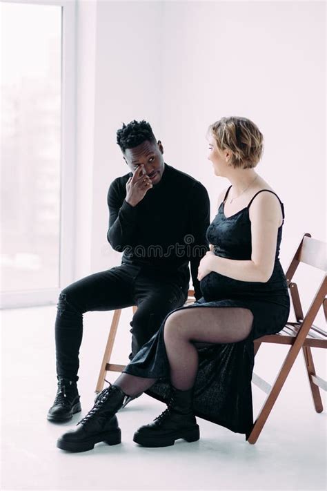 Young African Man And His Caucasian Pregnant Woman Sit On Chairs Stock