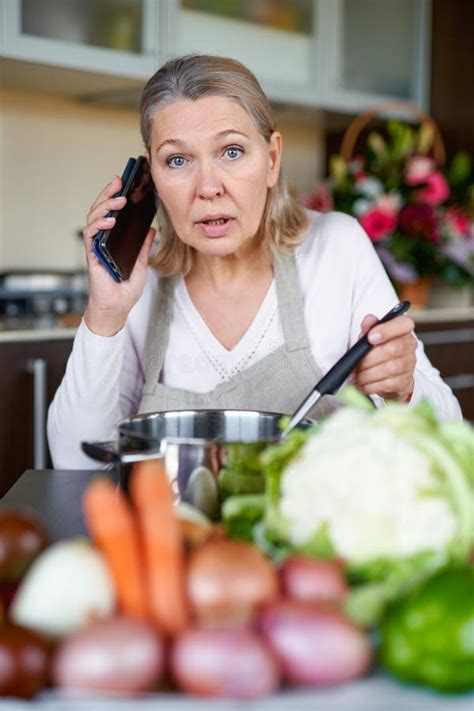 Mature Woman In Kitchen Preparing Food And Holding Smart Phone Stock