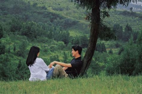 A Man And Woman Sitting Under A Tree On Top Of A Lush Green Hillside