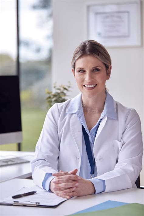 Portrait Of Smiling Female Doctor Or GP Wearing White Coat In Office