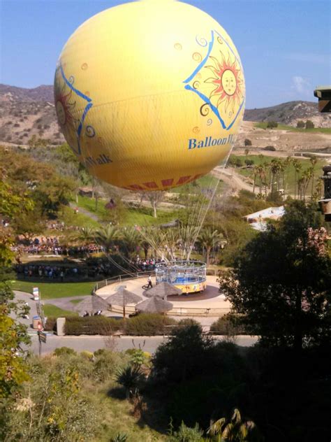 A Large Yellow Balloon Flying Over A Lush Green Hillside Next To A Park Filled With Trees