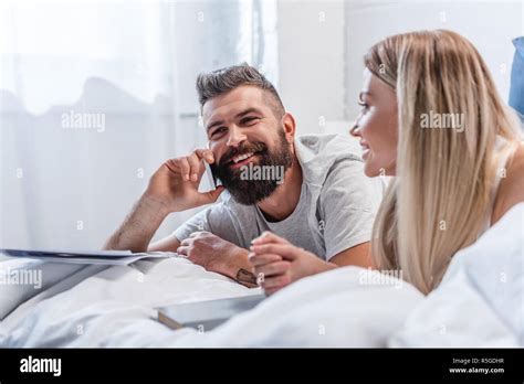 Bearded Man Talking On Smartphone While Blonde Girl Lying In Bed Stock