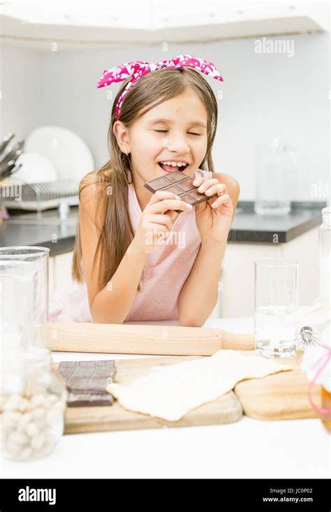 Portrait Of Smiling Cute Girl Biting Chocolate Bar On Kitchen Stock