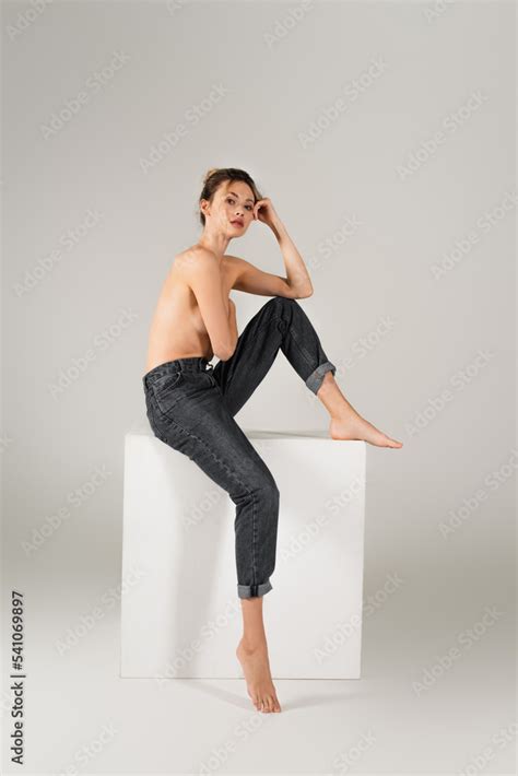 Sexy Half Nude And Barefoot Woman In Jeans Sitting On White Cube And