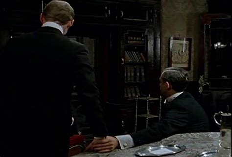 Two Men Sitting At A Table In Front Of A Bookcase With Books On It