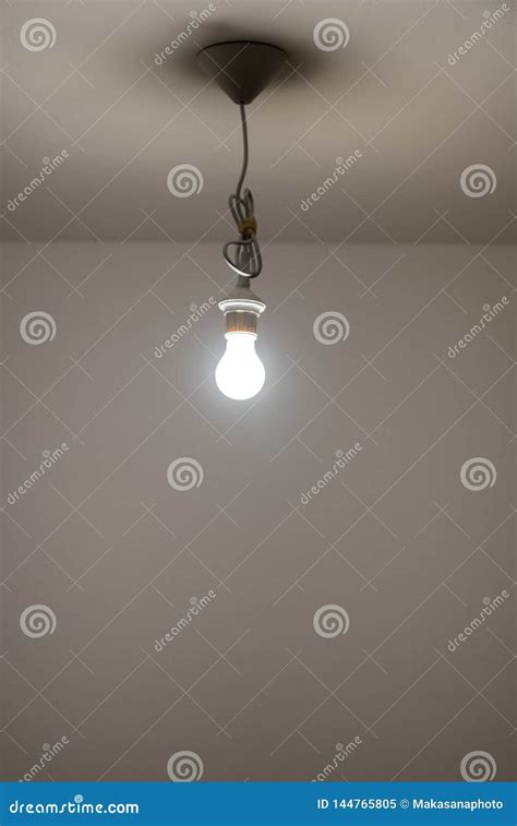 Naked Lit Light Bulb Hanging From The Ceiling Of A Dimly Lit Room Stock