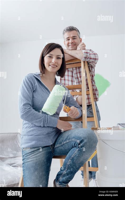 Happy Couple Renovating Their House And Painting A Room They Are Leaning On A Ladder And Posing